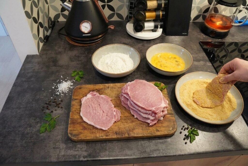 Stanowisko do panierowania schabowych na blacie kuchennym: rozbite mięso, miski z mąką, roztrzepanym jajkiem i bułką tartą