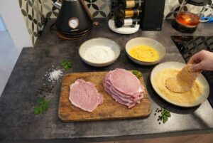 Stanowisko do panierowania schabowych na blacie kuchennym: rozbite mięso, miski z mąką, roztrzepanym jajkiem i bułką tartą